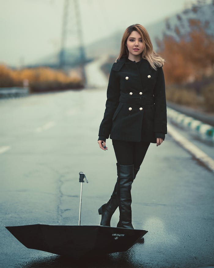 Stylish woman in black coat and boots stands with umbrella on a rainy street.